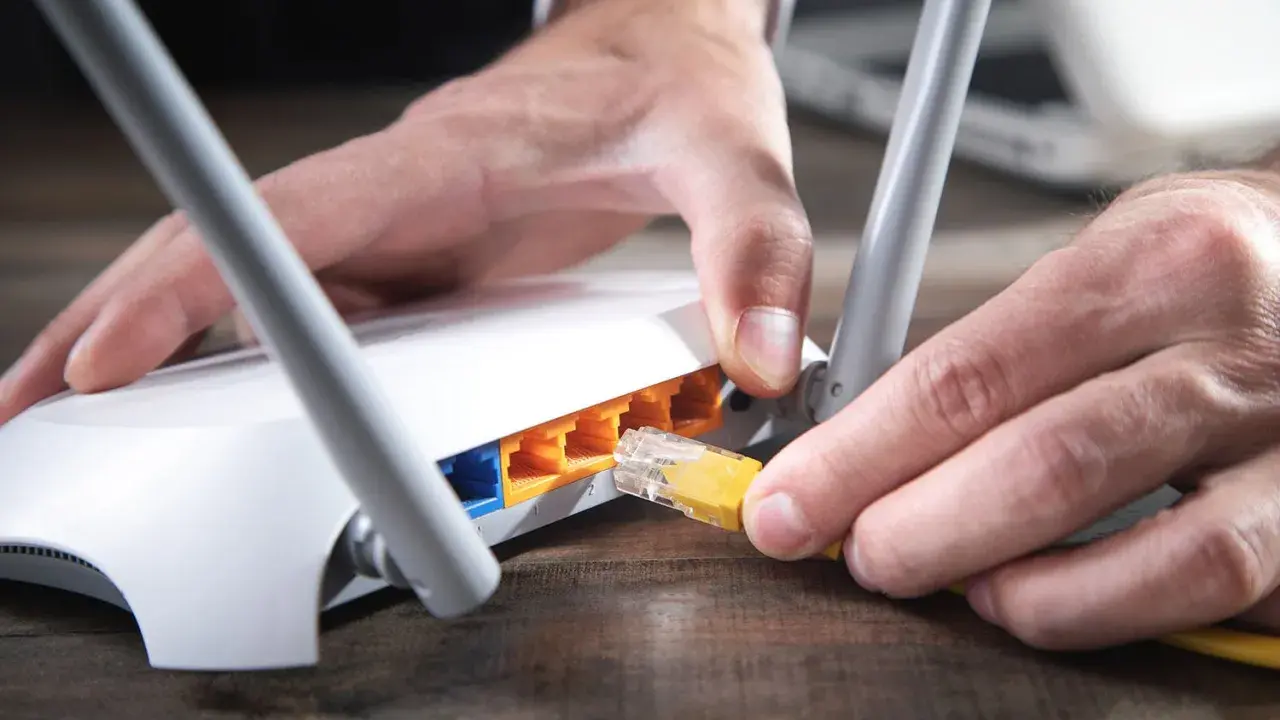 Händer kopplar in en gul nätverkskabel i en vit router, redo att byta router för bättre uppkoppling.