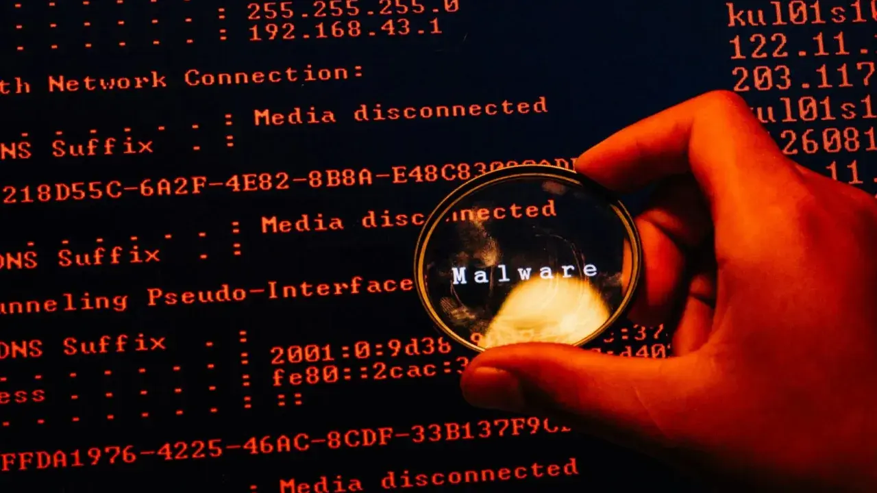 En hand håller ett förstoringsglas över en datorskärm. Förstoringsglaset visar ordet "Malware", vilket indikerar skadlig kod som döljer sig i systemets loggar.