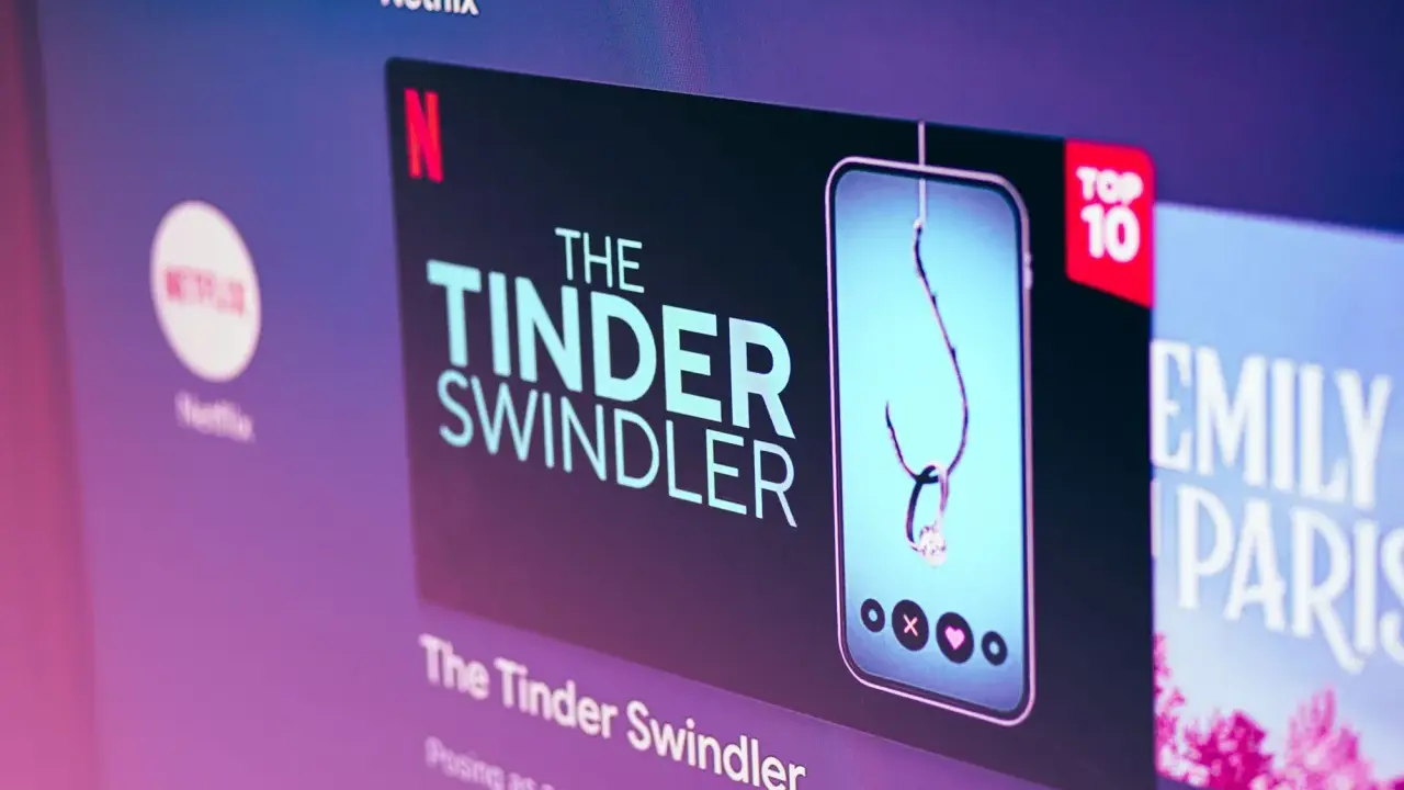 Netflix-skärmen visar "The Tinder Swindler", en dokumentär om tinder bilder på bedragare. En fiskekrok med en diamantring hänger i en mobil.