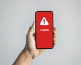 En hand håller en mobil med en röd skärm som visar en varningstriangel och texten "VIRUS". En påminnelse om hur vet man om man har virus.