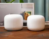 Två vita Google Nest Wifi-enheter står på ett träbord. En kartong med texten "Strong connection. Every direction." och Google-loggan syns i bakgrunden.