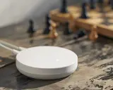 En vit, rund enhet, en dirigera hub, ligger på ett träbord bredvid ett schackbräde med pjäser.
