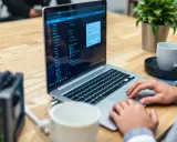 Person arbetar vid en laptop på ett träbord. En kaffekopp och en växt står bredvid. Dags att rensa datorn helt.