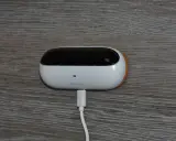 En vit, oval enhet med en svart topp, märkt "ambiclimate", är ansluten till en vit kabel. Den sitter på en träliknande yta och bidrar till ett uppkopplat hem.
