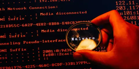 En hand håller ett förstoringsglas över en datorskärm. Förstoringsglaset visar ordet "Malware", vilket indikerar skadlig kod som döljer sig i systemets loggar.