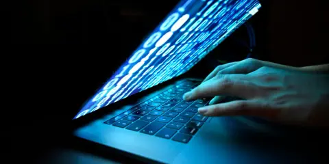 Händer på ett tangentbord, skärmen visar binär kod som flödar, en visuell representation av ett cyberattack idag.