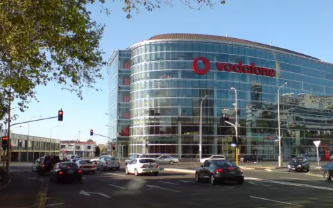 En modern glasbyggnad med stor Vodafone-logotyp, en livlig stadsgata med bilar och trafikljus. Vodafone Sverige syns tydligt.