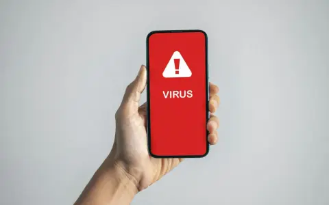 En hand håller en mobil med en röd skärm som visar en varningstriangel och texten "VIRUS". En påminnelse om hur vet man om man har virus.