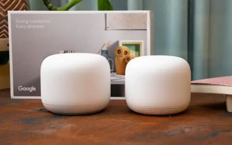 Två vita Google Nest Wifi-enheter står på ett träbord. En kartong med texten "Strong connection. Every direction." och Google-loggan syns i bakgrunden.