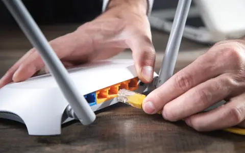 Händer kopplar in en gul nätverkskabel i en vit router, redo att byta router för bättre uppkoppling.