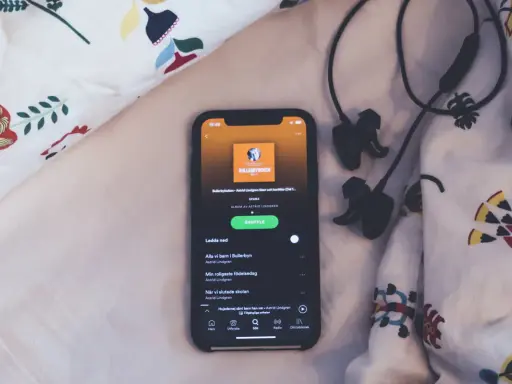 En mobil med Spotify-appen visar ljudboken "Bullerbyboken" av Astrid Lindgren, redo för insomningstimer. Hörlurar ligger bredvid.