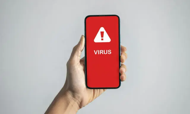 En hand håller en mobil med en röd skärm som visar en varningstriangel och texten "VIRUS". En påminnelse om hur vet man om man har virus.