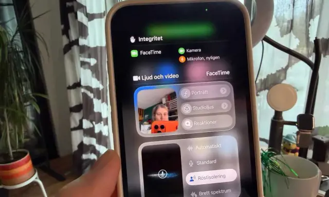 En person håller upp en iPhone som visar en skärm för gruppsamtal via FaceTime. Inställningar för ljud och video visas.