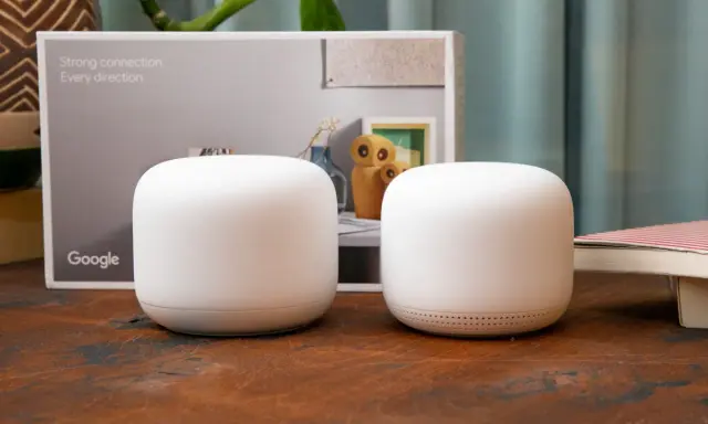 Två vita Google Nest Wifi-enheter står på ett träbord. En kartong med texten "Strong connection. Every direction." och Google-loggan syns i bakgrunden.