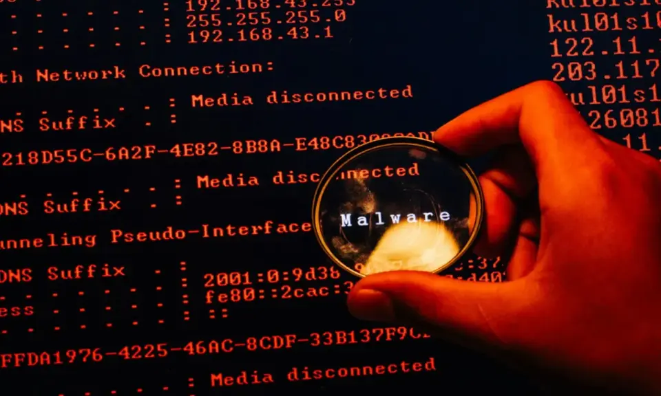 En hand håller ett förstoringsglas över en datorskärm. Förstoringsglaset visar ordet "Malware", vilket indikerar skadlig kod som döljer sig i systemets loggar.