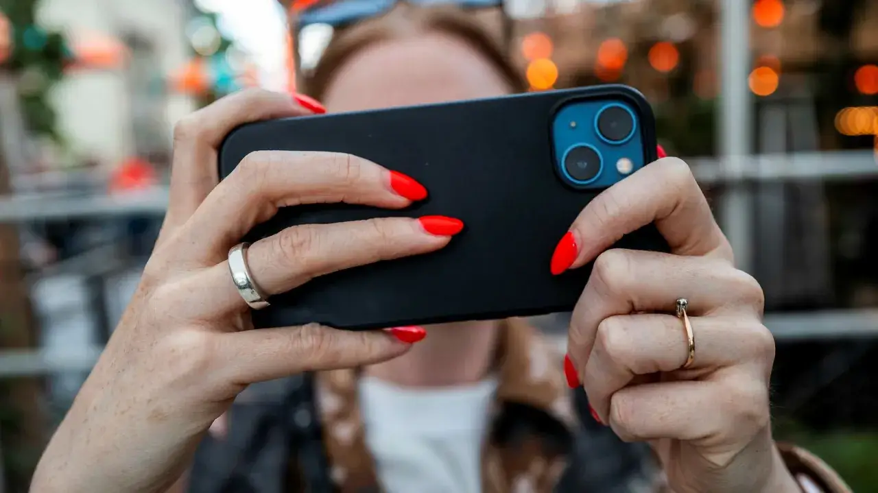 Händer med röda naglar håller en iPhone. Tänk på spionprogram iPhone när du delar din telefon.