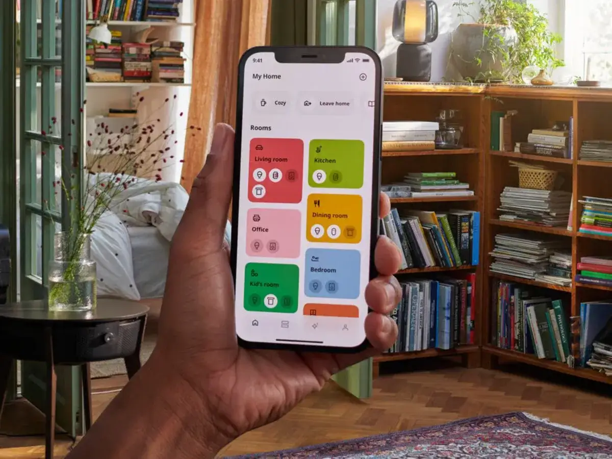 En hand håller en mobil med en app för att dirigera hubben i hemmet. Appen visar rum som kök och sovrum.