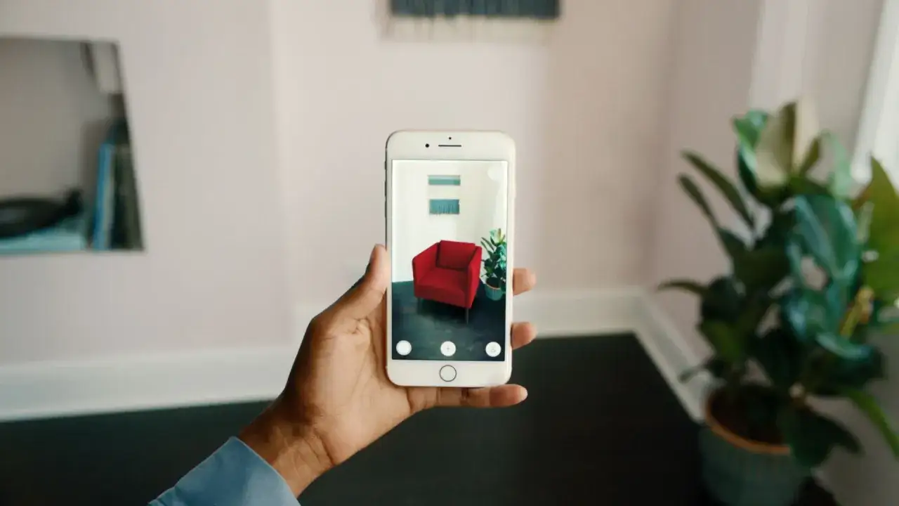En hand h&aring;ller en mobil med en r&ouml;d f&aring;t&ouml;lj och en v&auml;xt i AR. M&aring;ttband i mobilen hj&auml;lper till att visualisera m&ouml;bler i hemmet.