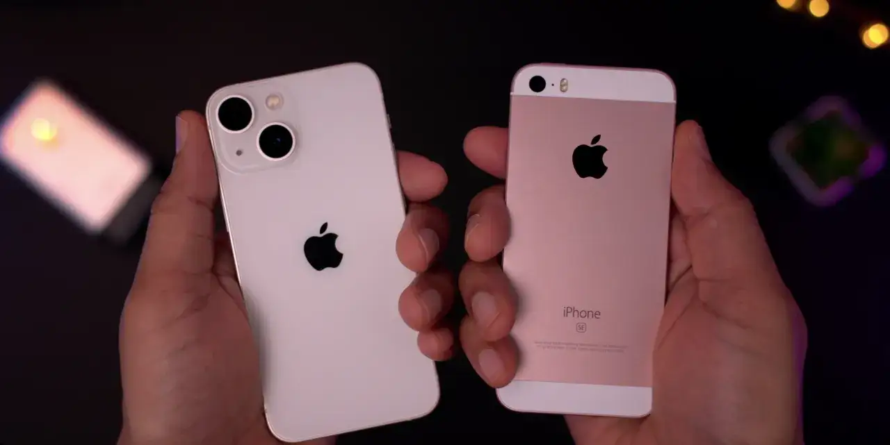 Två iPhones hålls upp, en vit iPhone 13 mini och en rosa iPhone SE. Jämför iphone 13 mini storlek med en äldre modell.