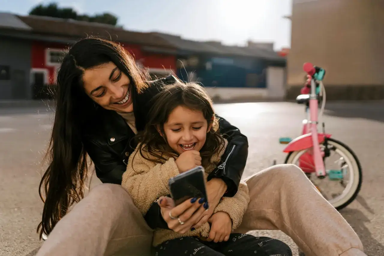 Mamma och dotter skrattar åt något på mobilen, kanske en rolig video i Telia appen. En rosa cykel syns i bakgrunden.