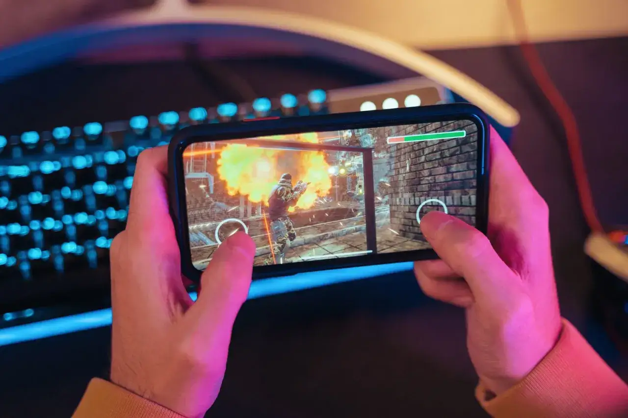 H&auml;nderna h&aring;ller en mobil med ett actionfyllt spel. En explosion syns p&aring; sk&auml;rmen, perfekt f&ouml;r roliga spel p&aring; mobilen.