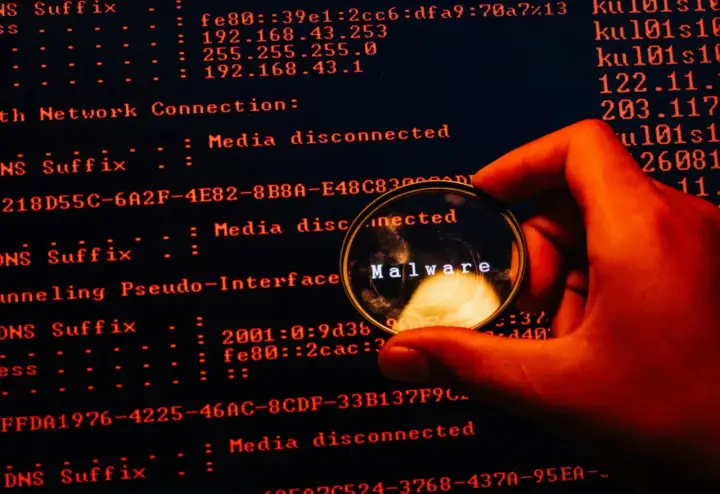 En hand håller ett förstoringsglas över en datorskärm. Förstoringsglaset visar ordet "Malware", vilket indikerar skadlig kod som döljer sig i systemets loggar.